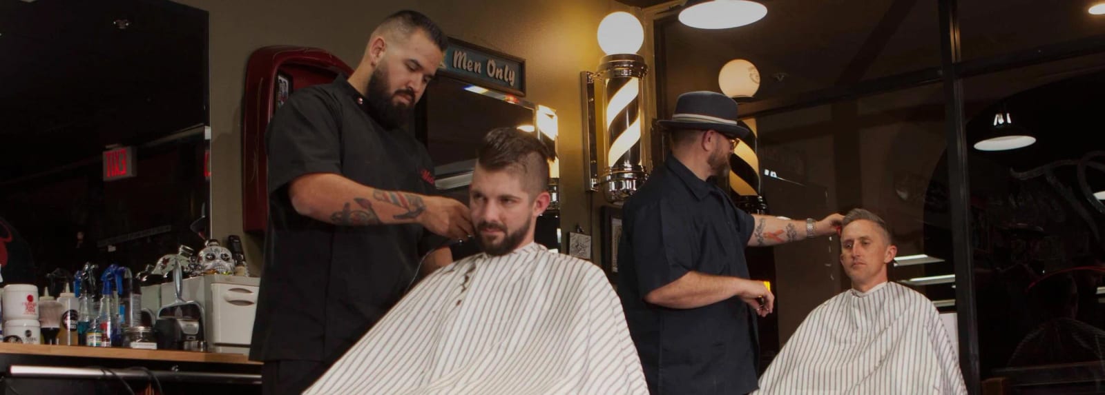 Two barbers cutting clients' hair beneath a Men Only sign and barber poles