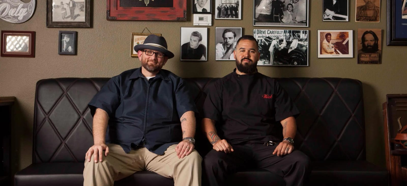 Two barbers seated on a bench in front of a wall of framed photos